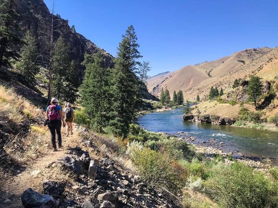 Two hikers with backpacks walking along a trail beside a river winding through the canyon
