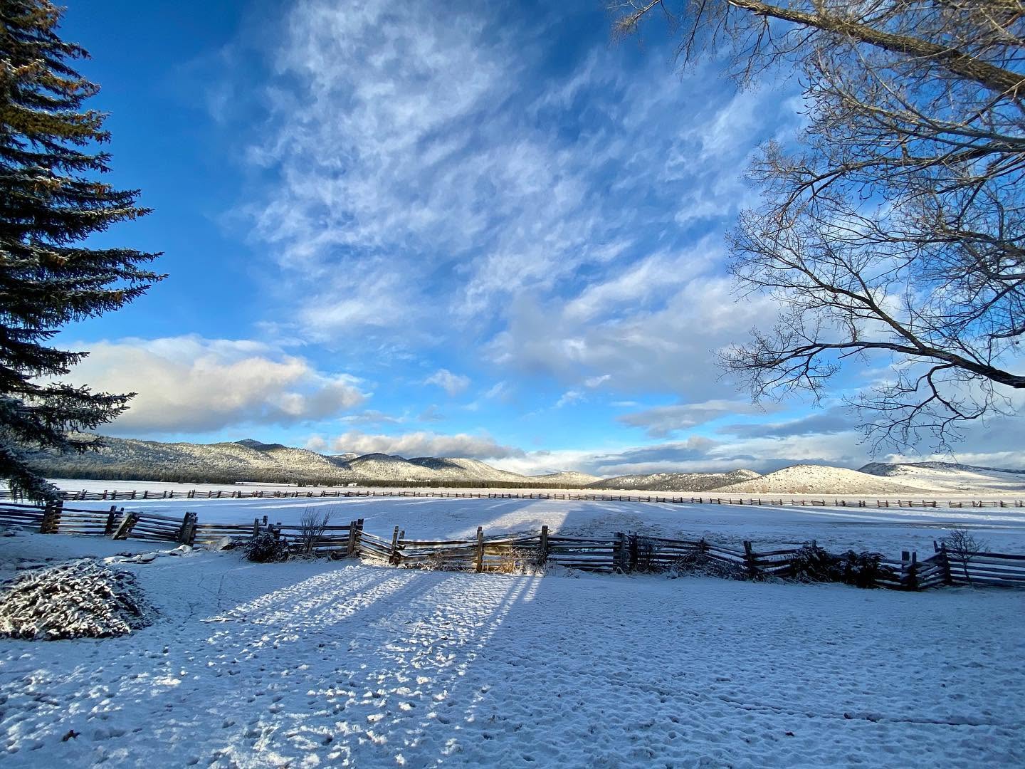 A snow-covered ranch pasture with a split-rail fence and mountains in the background
