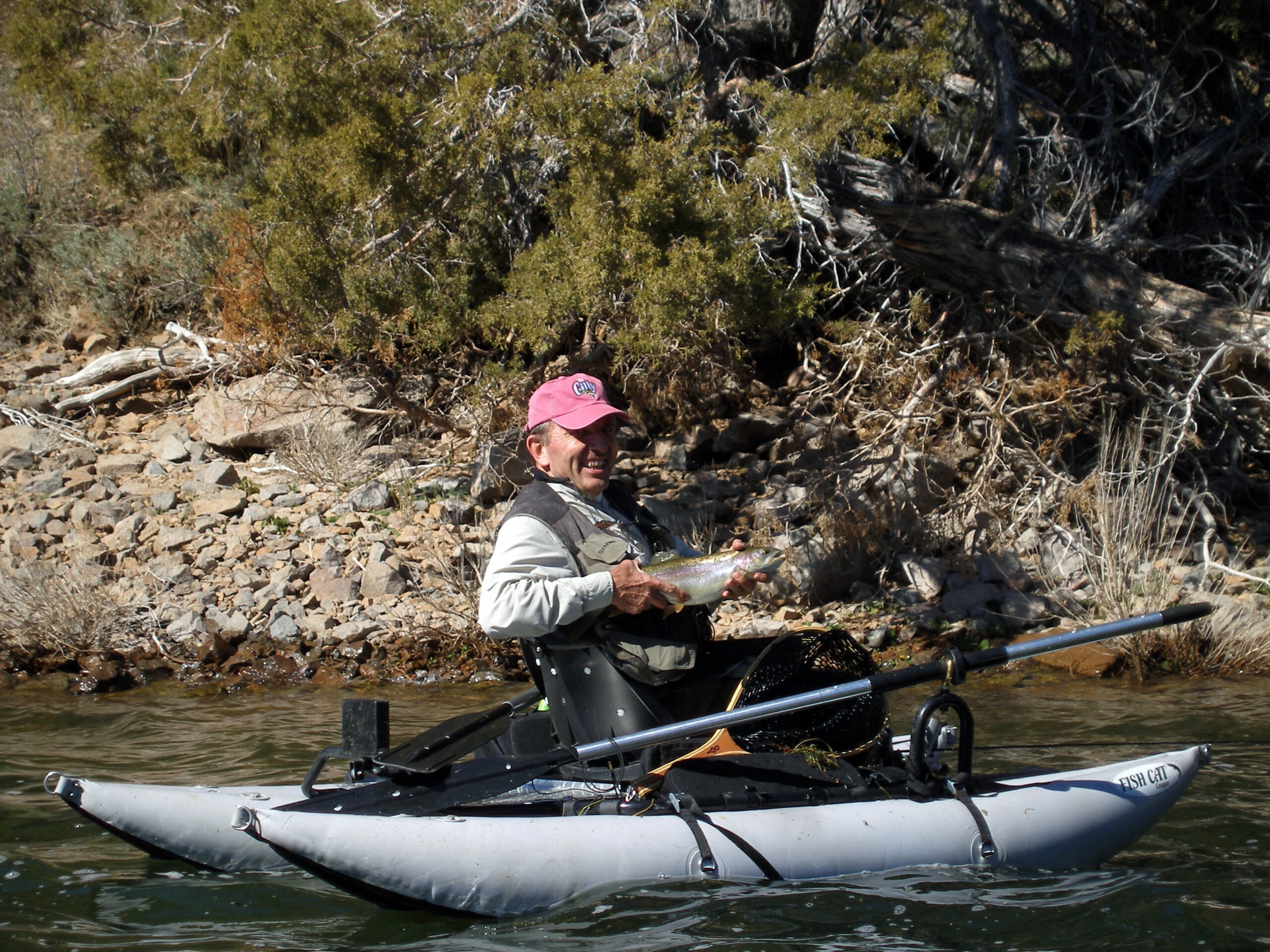 A man in a personal fishing pontoon boat smiling and holding a rainbow trout near the shore